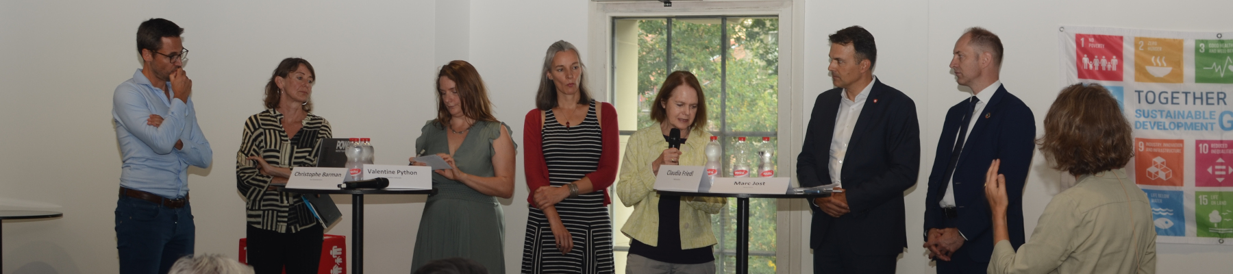 7 Personen stehen auf einem Podium. Im Hintergrund eine Fahne mit den SDG-Icons. Eine Frau steht vor den 7 Personen und spricht mit ihnen.